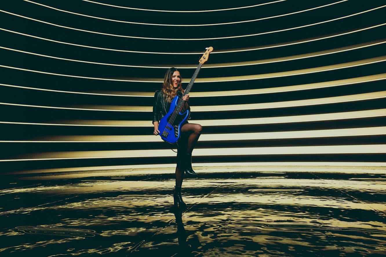 Woman playing blue electric bass guitar in a striped light tunnel, wearing black jacket, stockings, and heels, smiling at the camera.
