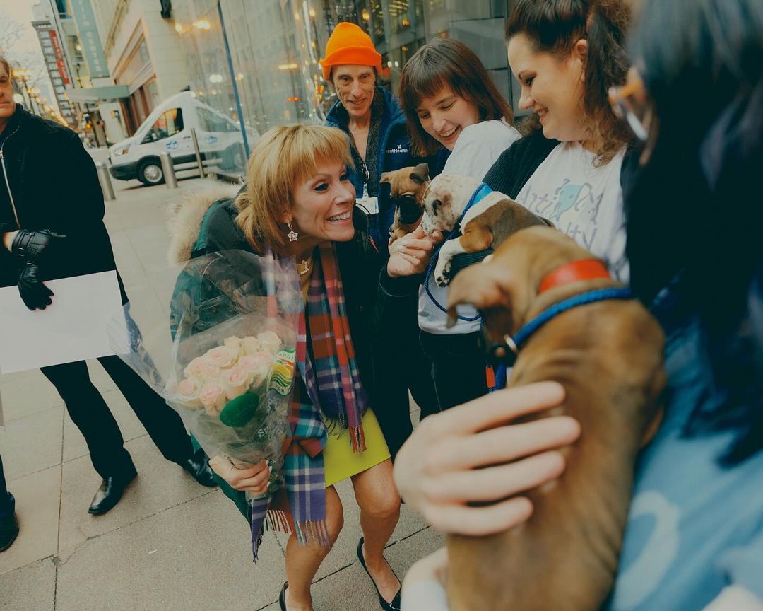 Happy woman in a yellow dress holds flowers outdoors surrounded by people with puppies on a city street with buildings behind.