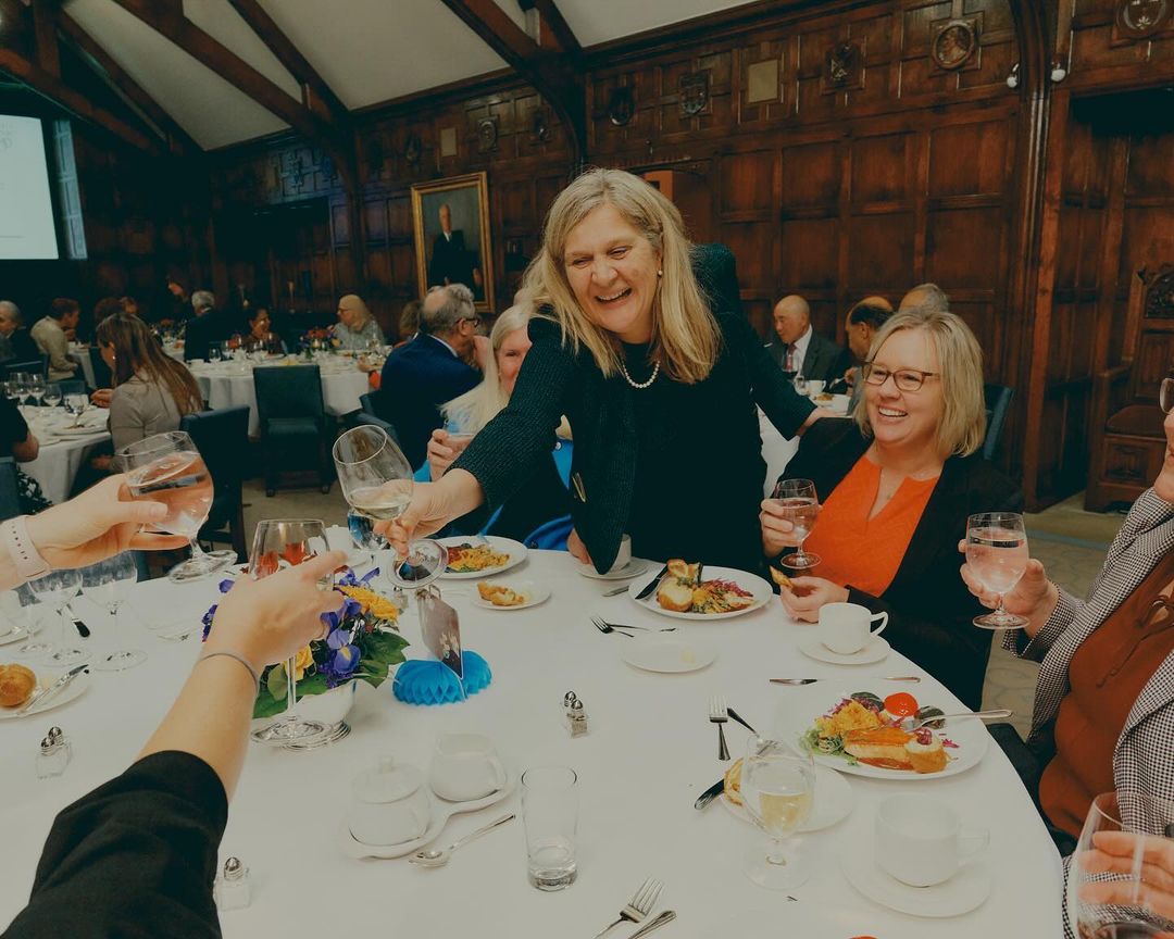 Group of people toasting with wine glasses at a banquet table inside a wood-paneled room with a portrait hanging on the wall.