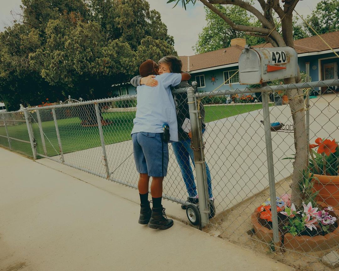 Two people embracing outside a home with a mailbox numbered 420. One person wears a postal worker uniform with blue shorts and black boots.
