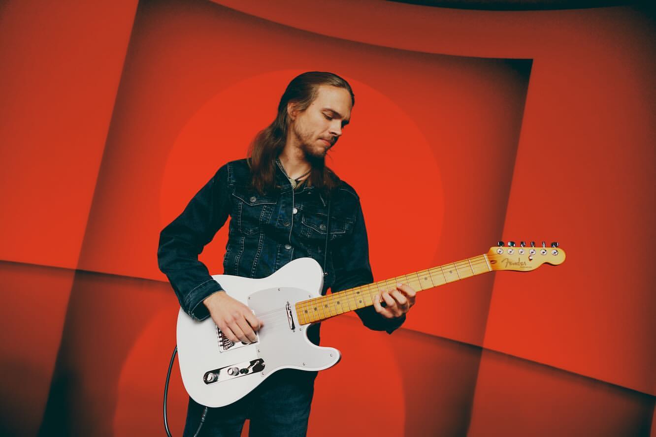 Adult male musician playing a white electric guitar with a red abstract background in an indoor music studio setting. 