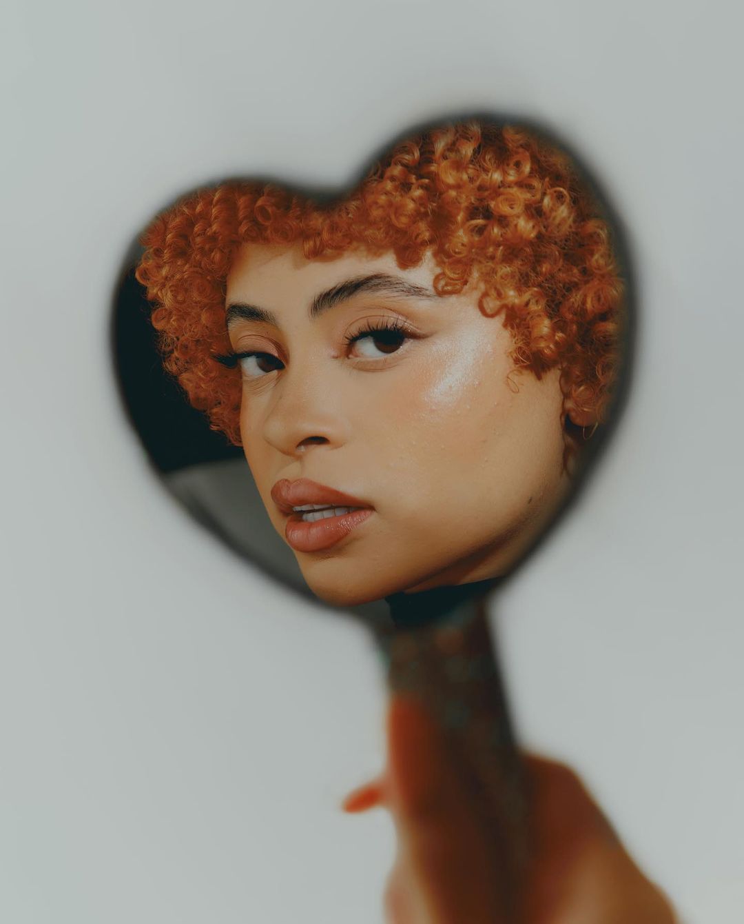 Close-up studio portrait of an adult female with auburn curly hair, holding up a heart-shaped mirror reflecting her face against a white background.