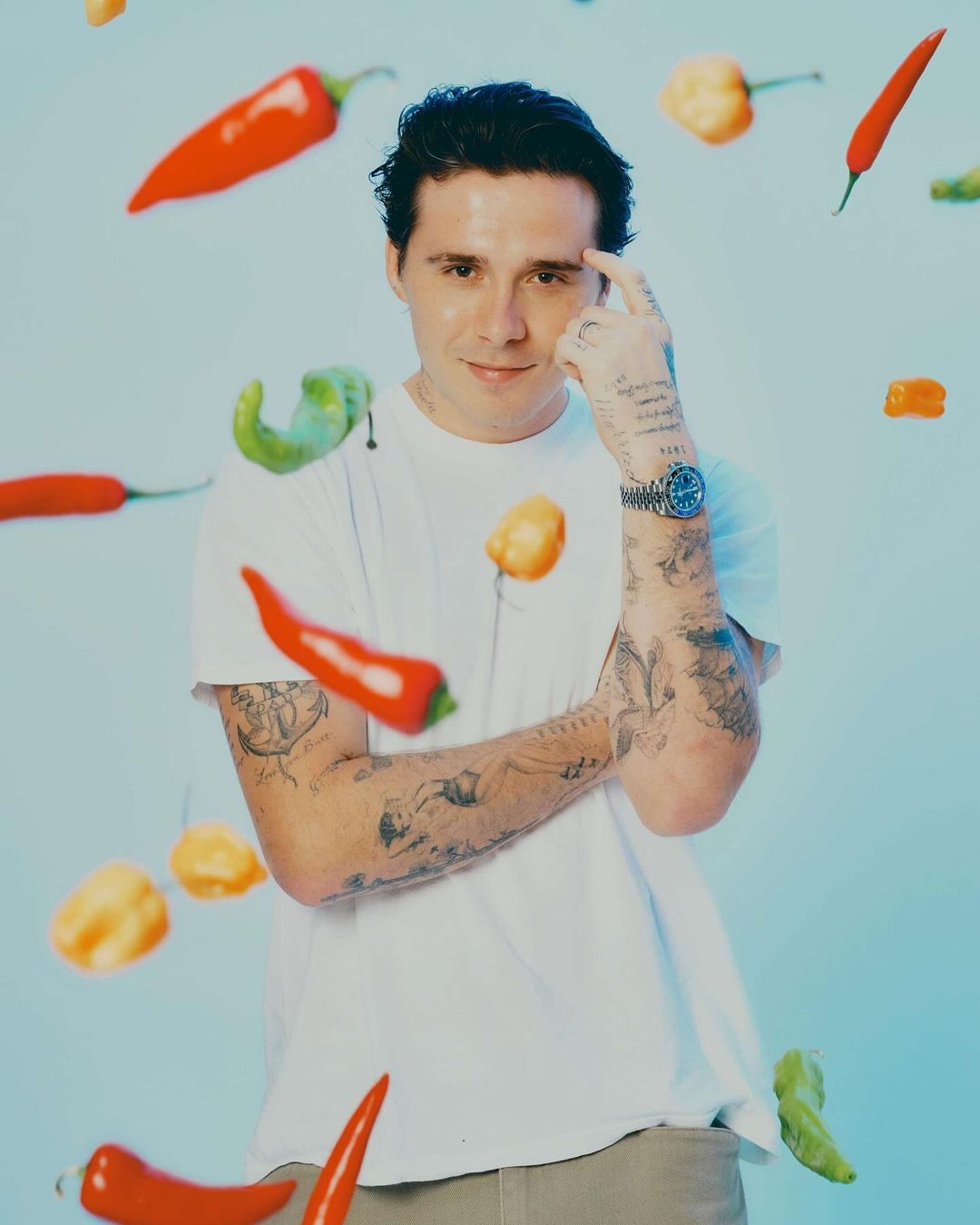 Portrait of a tattooed adult male in a white t-shirt surrounded by red, yellow, and green hot peppers against a pale blue background.