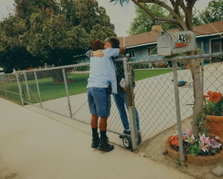 Two people hugging over a chain link fence in front of a residential house with a mailbox reading 420.