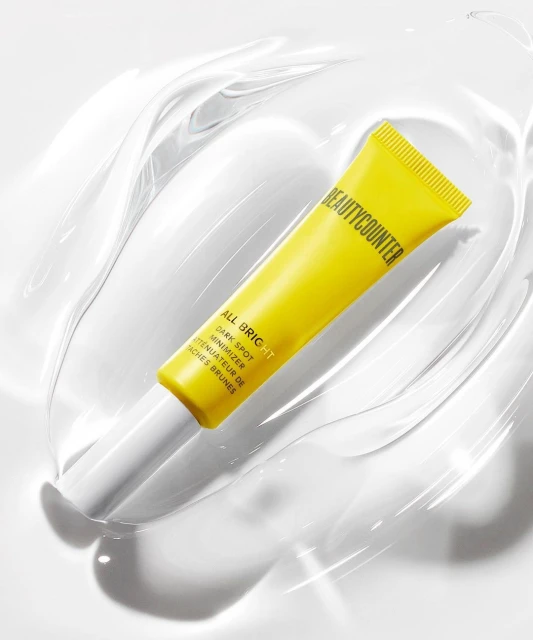 Close-up of yellow Beautycounter All Bright Dark Spot Minimizer tube with white accents, sitting on a clear, glossy, fluid-like surface.