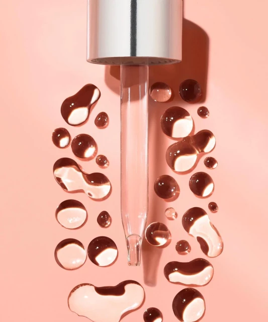 Close-up shot of a silver and clear cosmetic dropper surrounded by clear serum drops on a light pink background creating an eye-catching beauty image.