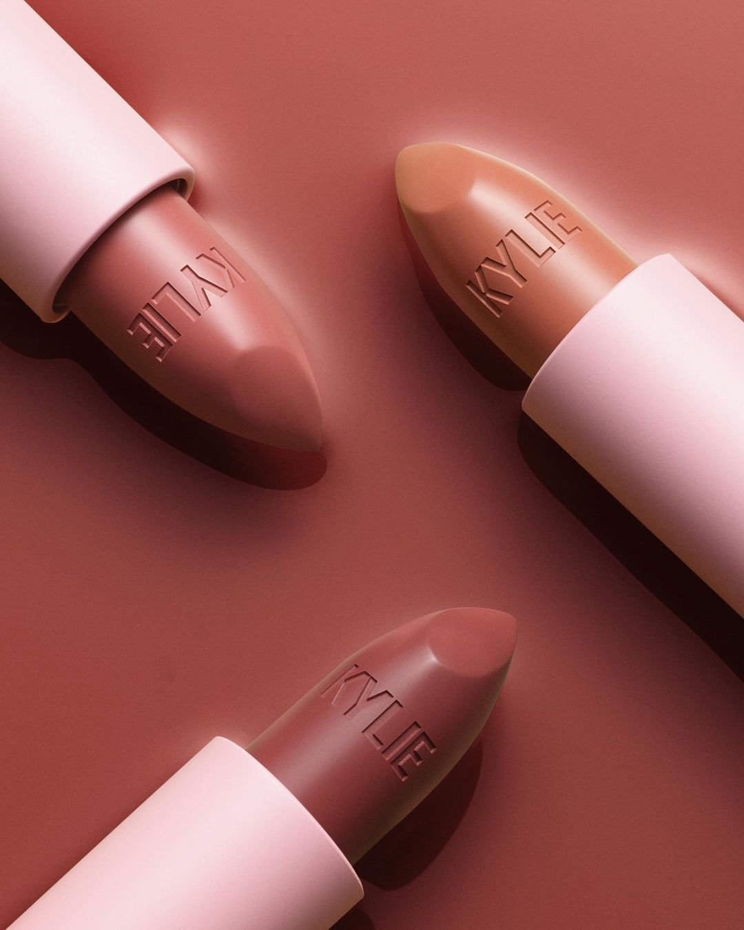 Three Kylie lipsticks in different shades of brown and nude, displayed on a matching brown background in a beauty product still life.