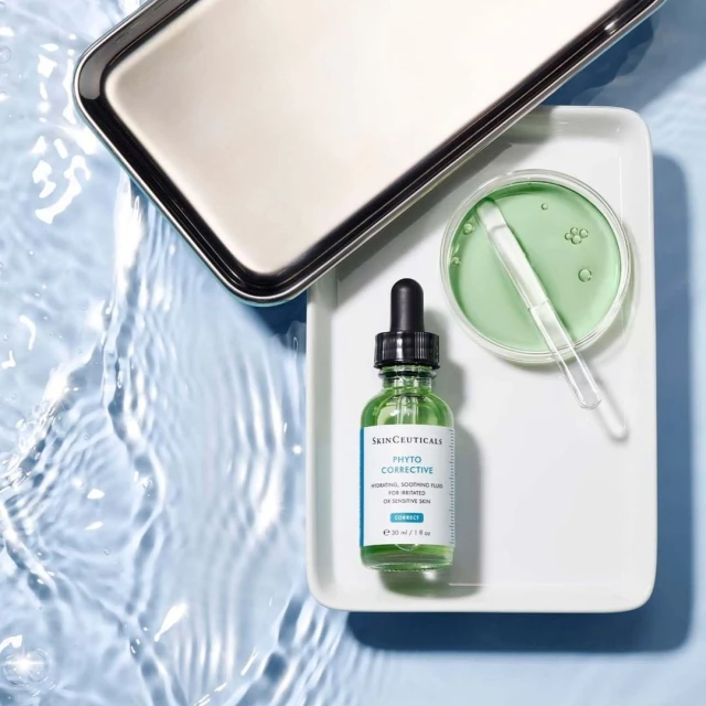 Still life of SkinCeuticals Phyto Corrective serum bottle with dropper, petri dish, and metal tray on a white surface with water ripples.