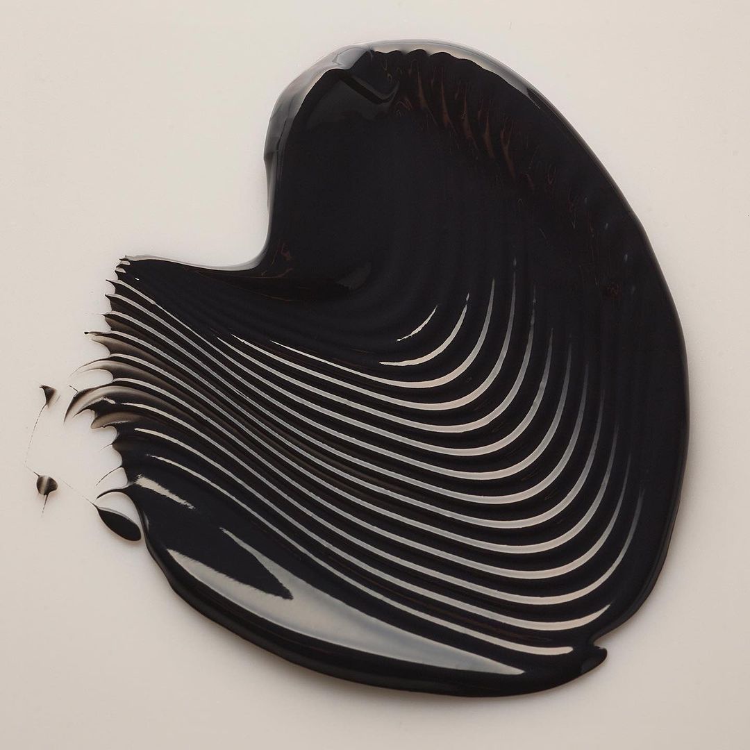 Dark, glossy liquid with a ribbed texture is spread across a pale background, creating an abstract pattern with a smooth and shiny surface.