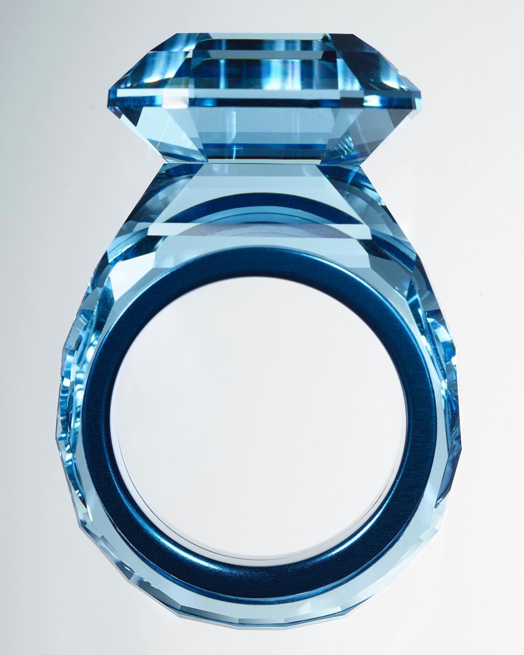 Close-up studio shot of a large clear light-blue decorative ring made of crystal or glass with geometric facets, on a gradient white background.