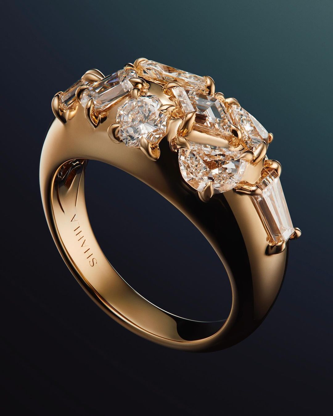 Elevated view of an ornate gold ring set with various cut diamonds against a deep teal backdrop in a jewelry product shot.