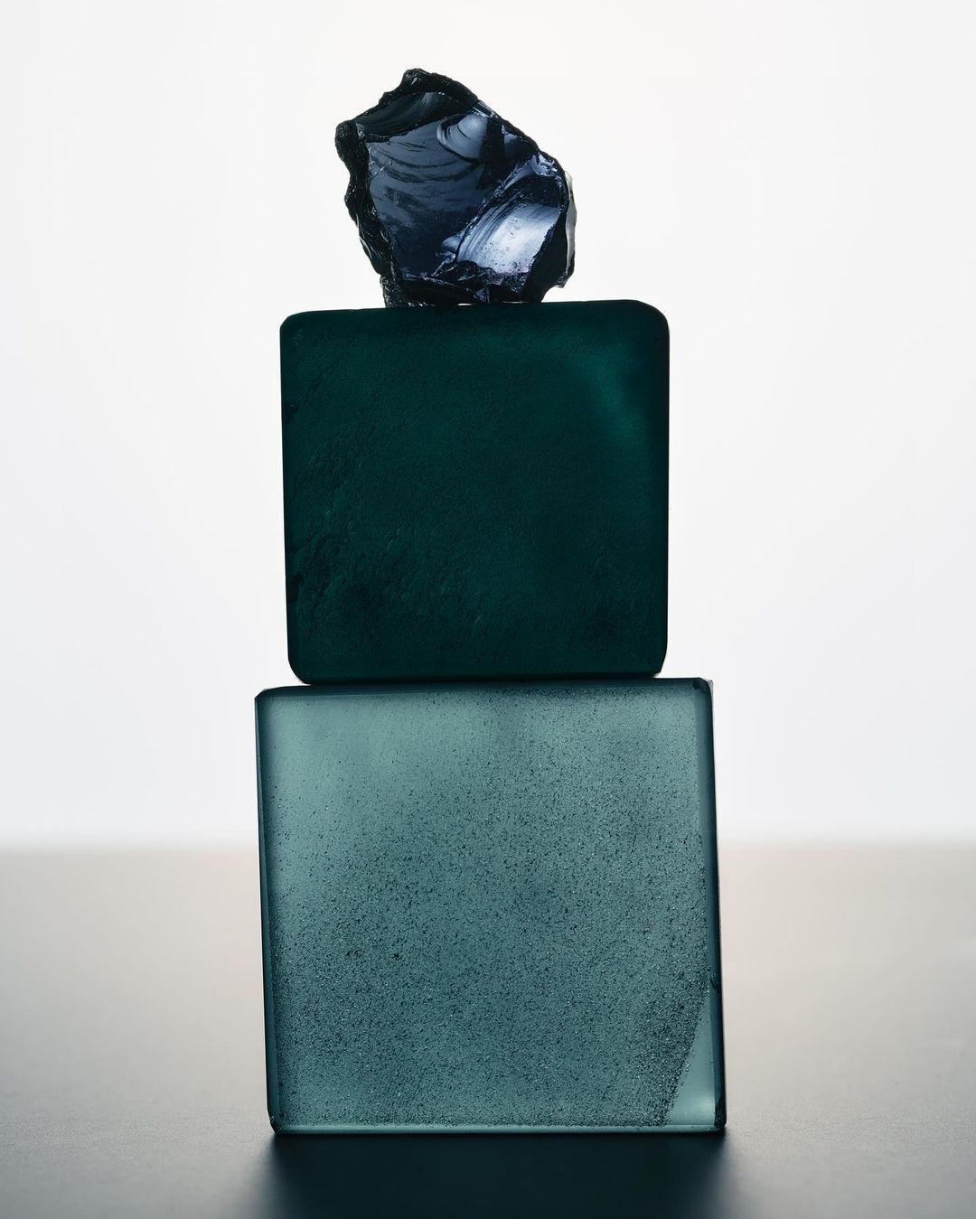 Still life photograph featuring two square teal-colored glass blocks stacked, topped with a dark, irregularly-shaped glass piece against a white backdrop.