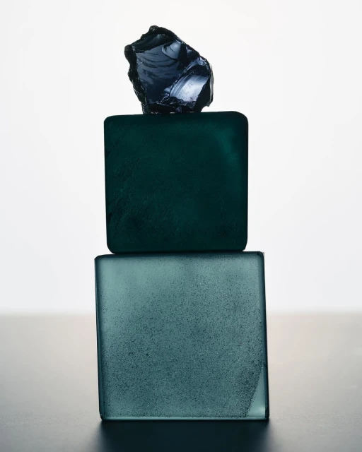 Still life photograph featuring two square teal-colored glass blocks stacked, topped with a dark, irregularly-shaped glass piece against a white backdrop.