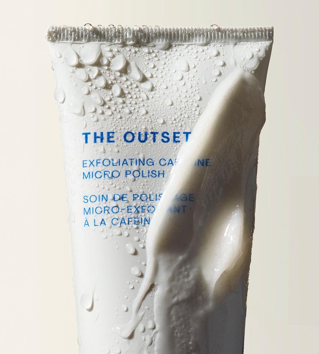 Close-up of white cosmetic tube with 'THE OUTSET' brand name, water droplets, and smear of cream. Beauty and skincare product.