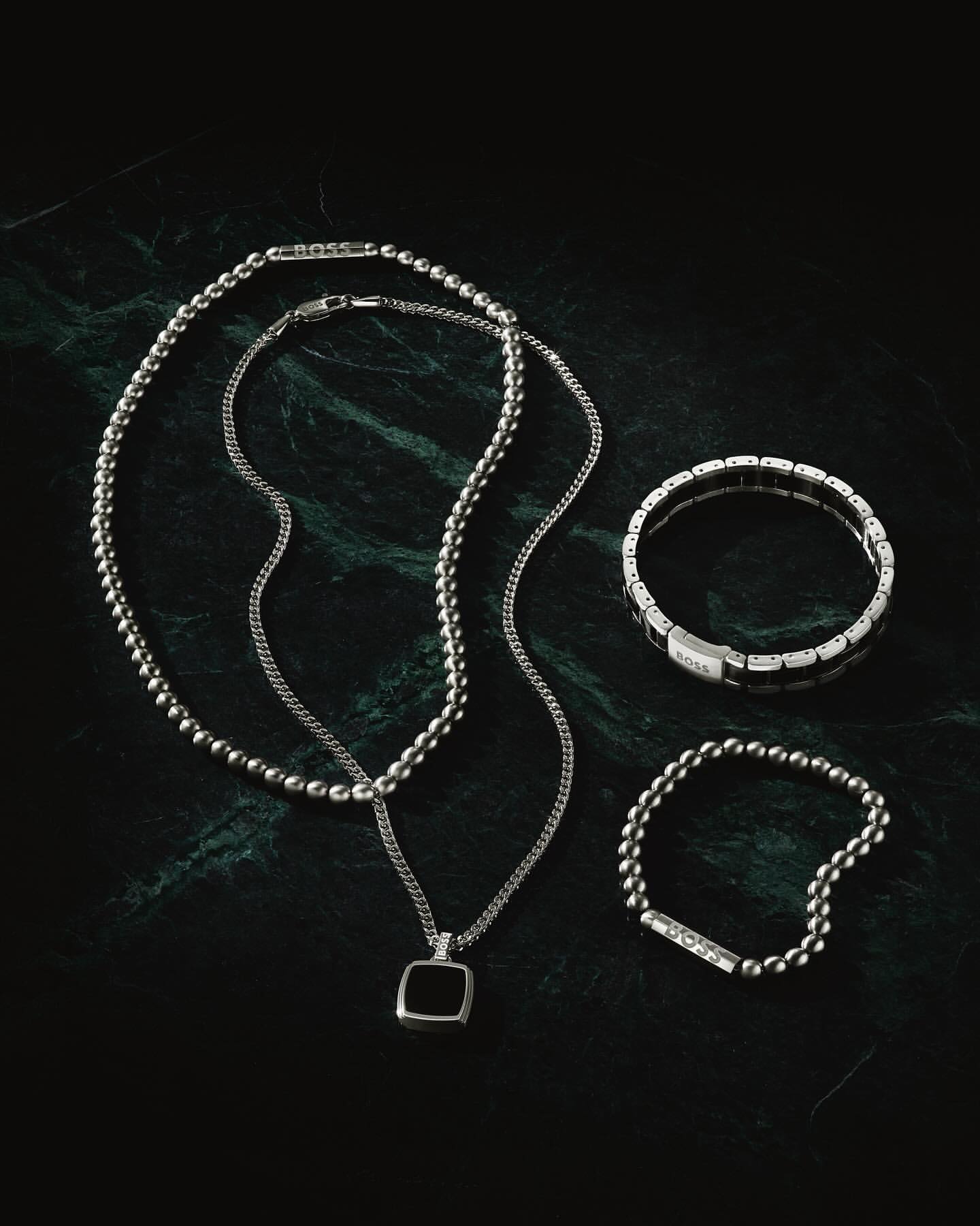 Close-up product shot on dark green marble featuring a ball chain necklace, pendant necklace, beaded bracelet, and panel bracelet.