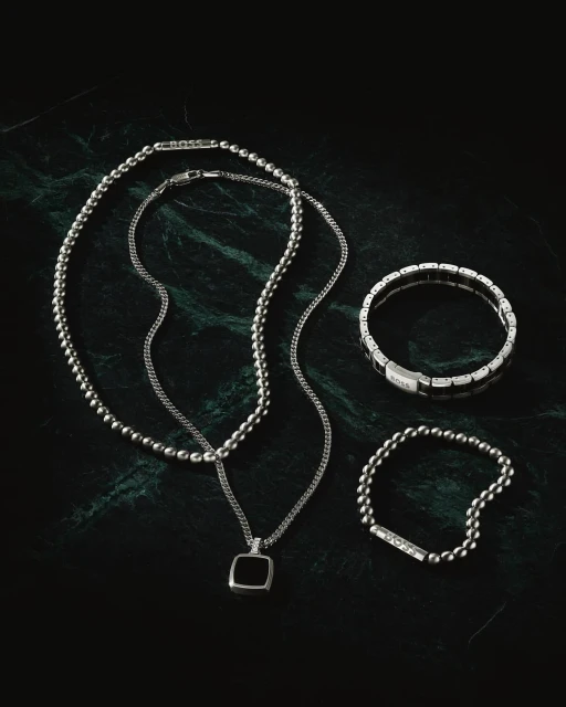 Close-up product shot on dark green marble featuring a ball chain necklace, pendant necklace, beaded bracelet, and panel bracelet.