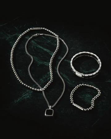 Close-up product shot on dark green marble featuring a ball chain necklace, pendant necklace, beaded bracelet, and panel bracelet.