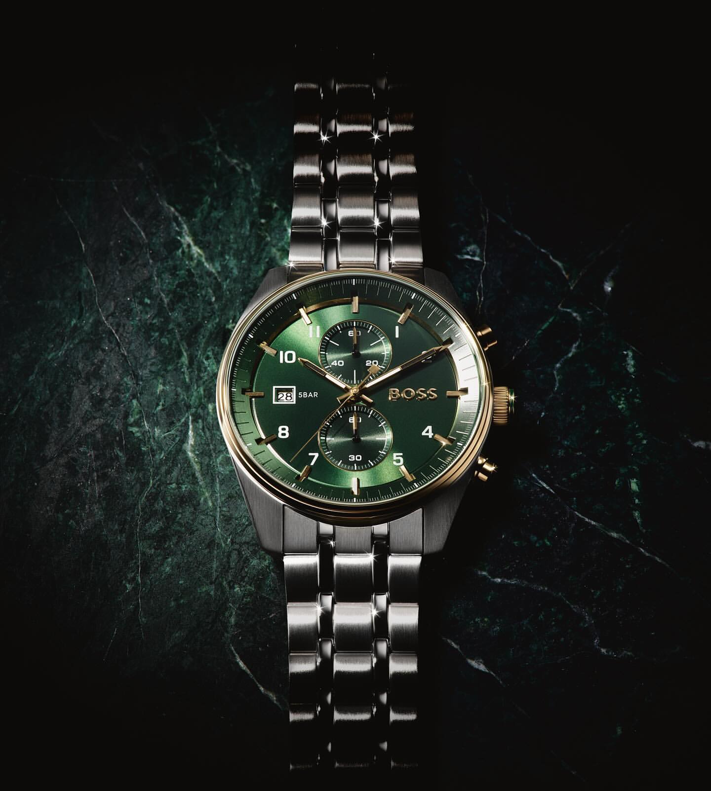 Close-up studio shot of a Hugo Boss wristwatch with a silver band and a green dial on a dark green, veined marble surface.