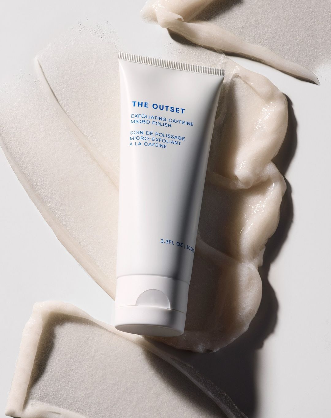 Product shot of white tube of The Outset exfoliating caffeine micro polish surrounded by smears of the same product on a white surface.