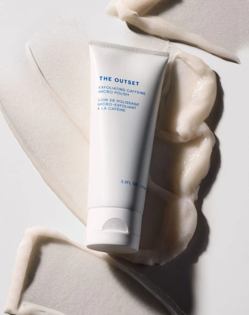 Product shot of white tube of The Outset exfoliating caffeine micro polish surrounded by smears of the same product on a white surface.
