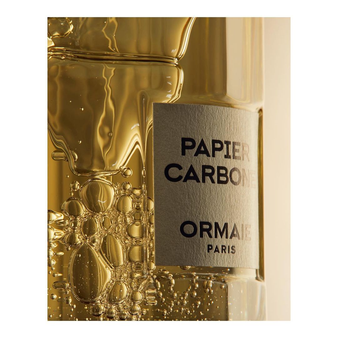 Close-up studio shot of a transparent glass bottle filled with gold liquid and bubbles; gray label reads: Papier Carbone, Ormaie Paris.