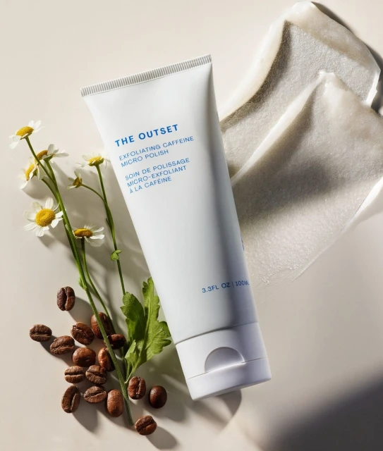 White tube of The Outset exfoliating caffeine micro polish surrounded by coffee beans and chamomile flowers on a light beige surface.