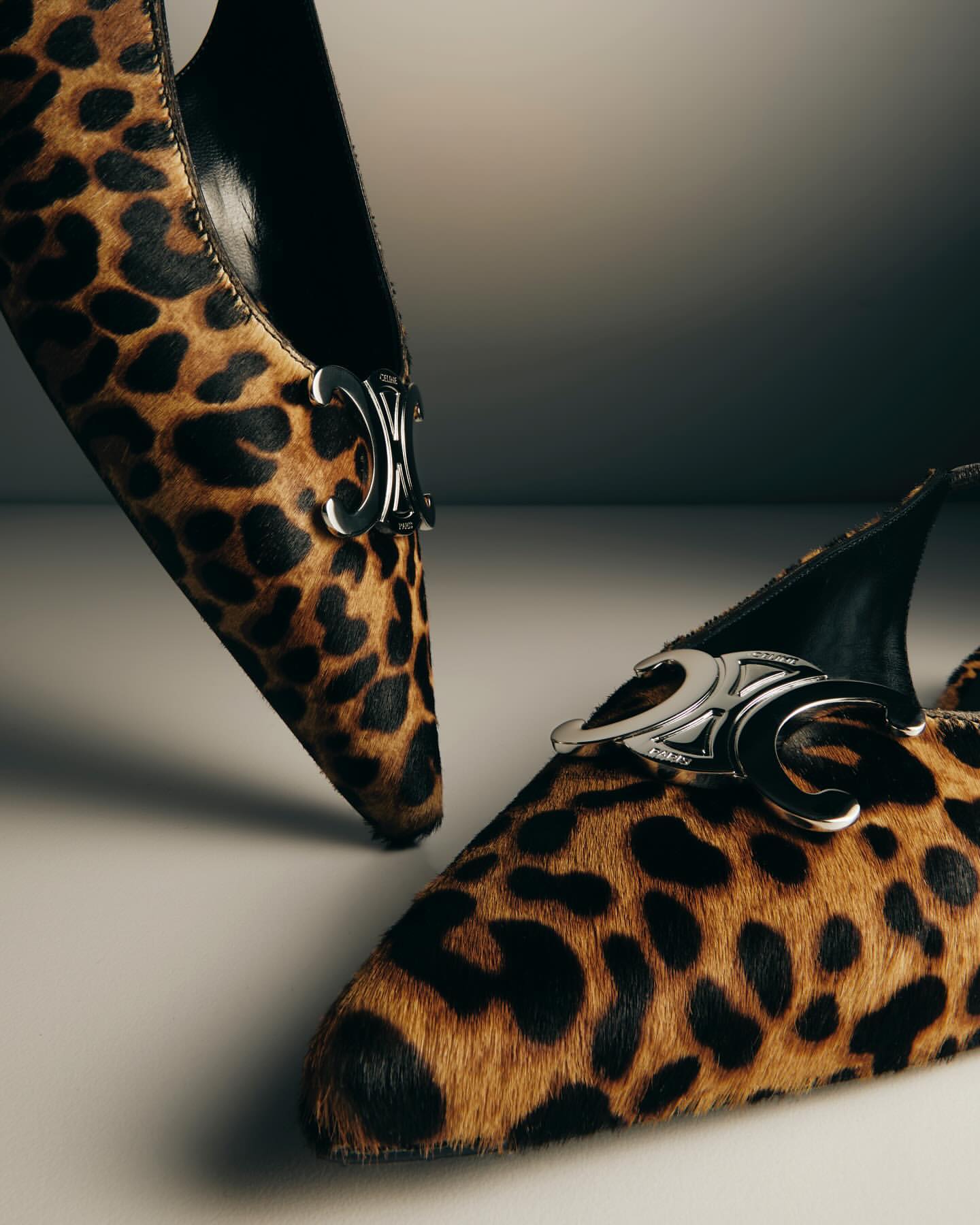 Two tan and black leopard print heeled pumps with silver hardware sit on a white surface against a white and gray background.