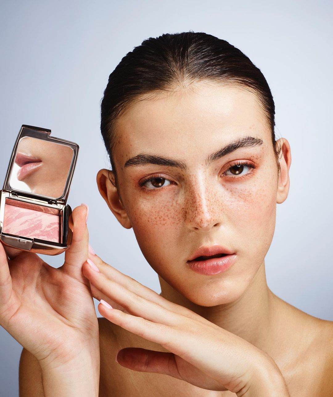 Beauty portrait of a fair-skinned woman holding a compact of blush, showcasing makeup application against a light background in studio setting.