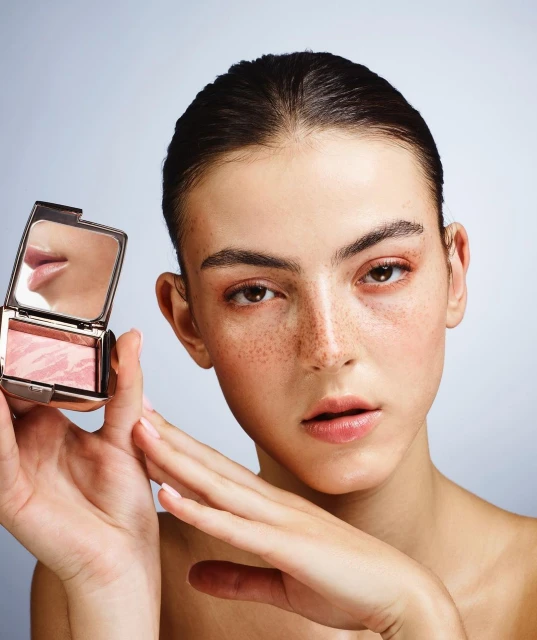 Beauty portrait of a fair-skinned woman holding a compact of blush, showcasing makeup application against a light background in studio setting.