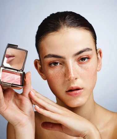 Beauty portrait of a fair-skinned woman holding a compact of blush, showcasing makeup application against a light background in studio setting.