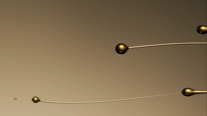 Macro shot of three small golden liquid drops suspended on thin wires against a blurred golden background; clean minimalist design.