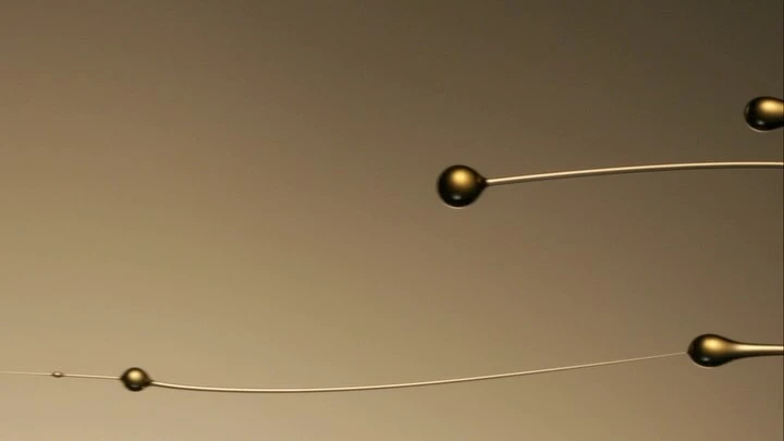 Macro shot of three small golden liquid drops suspended on thin wires against a blurred golden background; clean minimalist design.