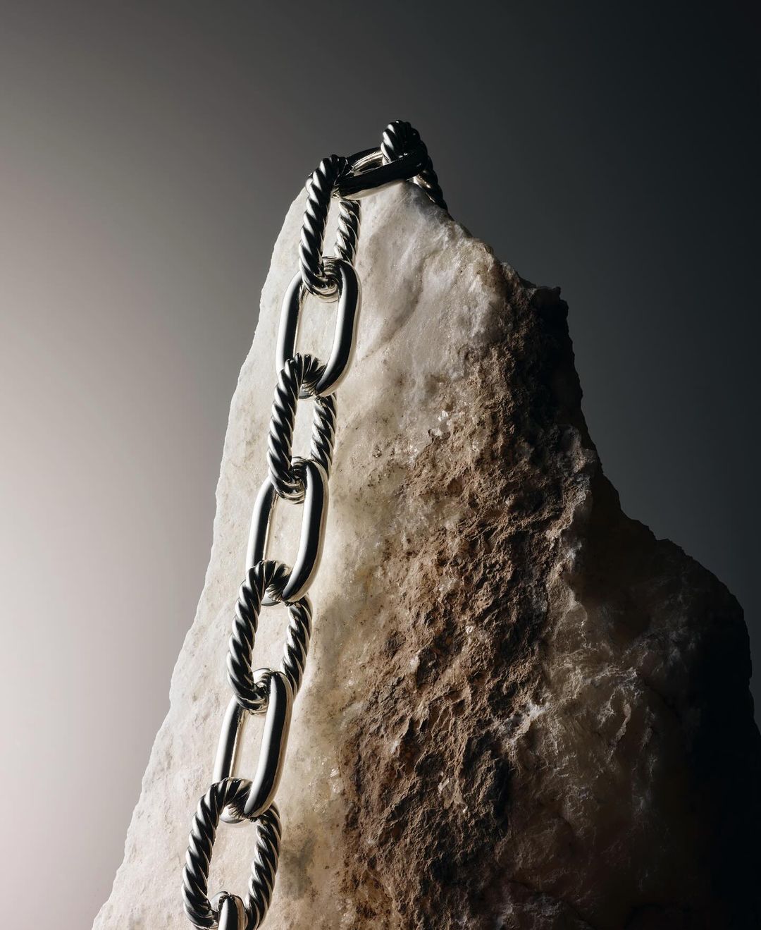 Silver cable chain necklace draped over a rough light-colored stone against a dark gradient background
