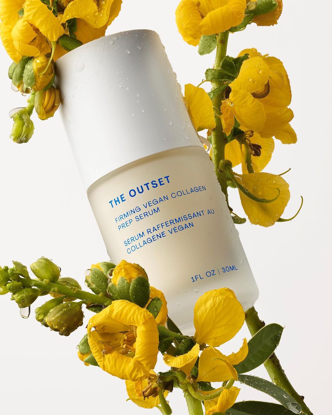 Serum bottle from The Outset, surrounded by yellow flowers, showcasing vegan collagen prep in a beauty product shot on white.