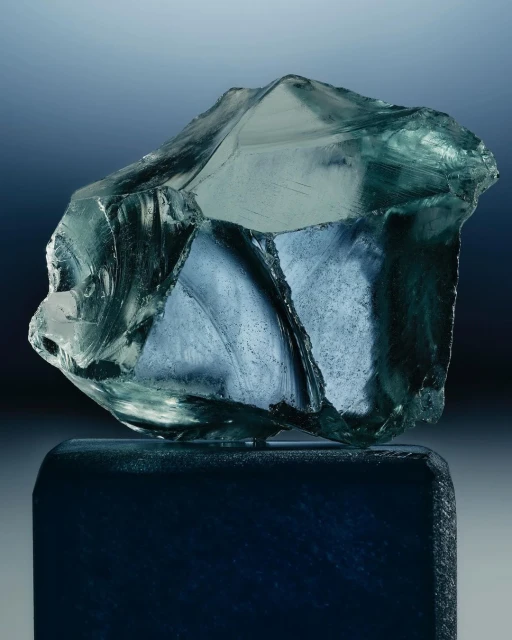 Translucent block of blue-green mineral glass sitting atop a dark blue, rectangular pedestal against a graduated blue background.