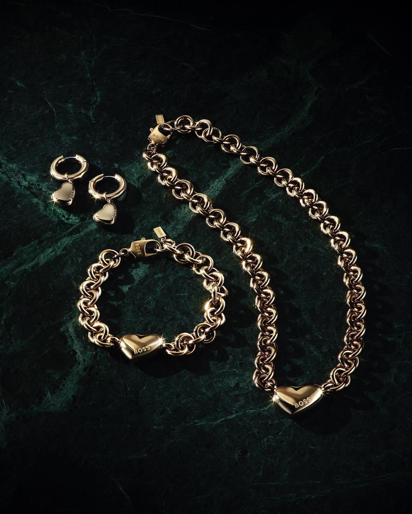 Gold-toned 'BOSS' jewelry set on dark green marbled surface, including necklace, bracelet, and hoop earrings with heart pendants.