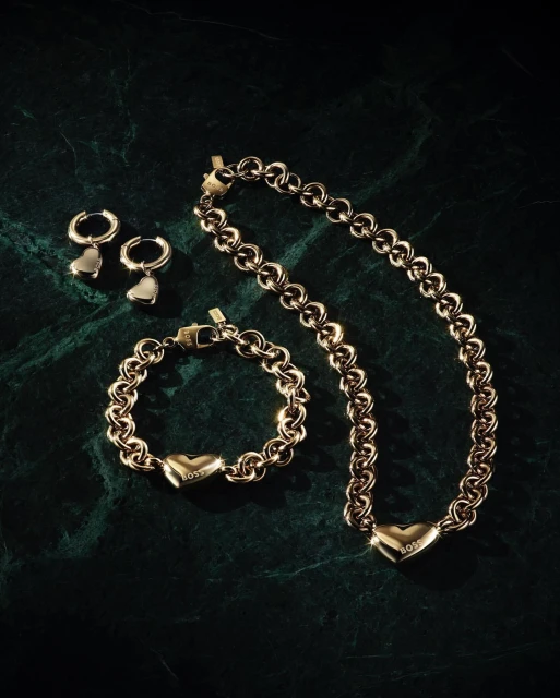 Gold-toned 'BOSS' jewelry set on dark green marbled surface, including necklace, bracelet, and hoop earrings with heart pendants.