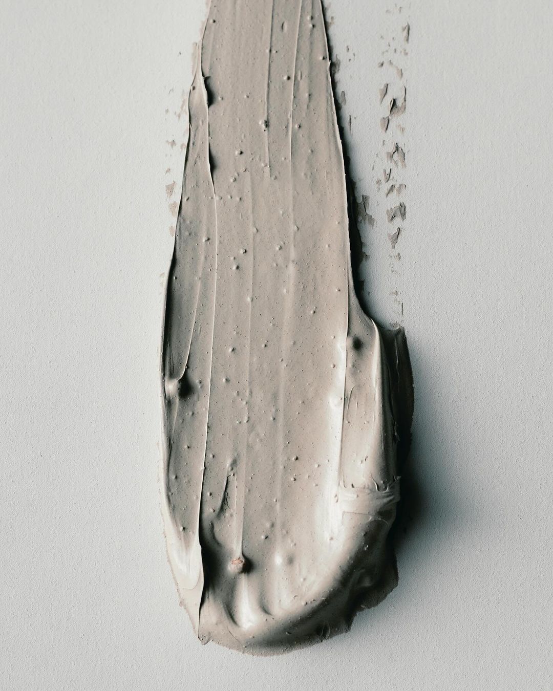 Close-up shot of a smear of greyish-beige cosmetic cream on a white surface, showcasing its texture and color.
