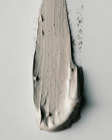 Close-up shot of a smear of greyish-beige cosmetic cream on a white surface, showcasing its texture and color.