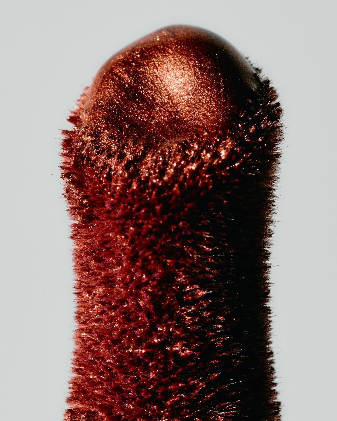 Close-up of a copper-colored cosmetic product stick. The end is rounded, with a grainy texture and a reflective sheen against a light background.