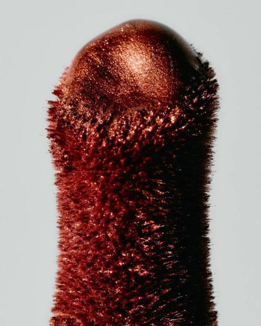 Close-up of a copper-colored cosmetic product stick. The end is rounded, with a grainy texture and a reflective sheen against a light background.