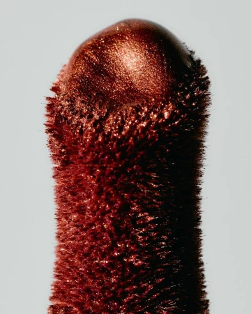 Close-up of a copper-colored cosmetic product stick. The end is rounded, with a grainy texture and a reflective sheen against a light background.