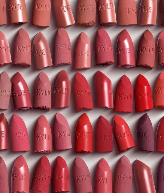 Close-up of Kylie lipsticks in various red and nude shades arranged in a grid pattern; Kylie logo visible on each cosmetic product.