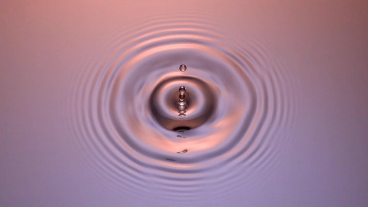 Macro photography of a water droplet causing ripples in a pool of liquid with soft pink and purple gradient lighting.
