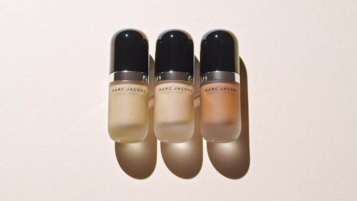 Three Marc Jacobs foundation bottles with varying shades sit closely together on a soft off-white surface, casting distinct shadows.