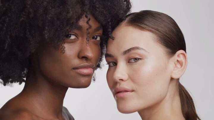 Close-up portrait of two diverse female models with natural makeup posing closely against a neutral white background.