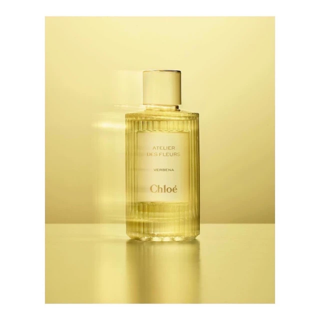 Elegant still life showcases a glass bottle of Chloé Atelier des Fleurs Verbena perfume against a gradient golden background.
