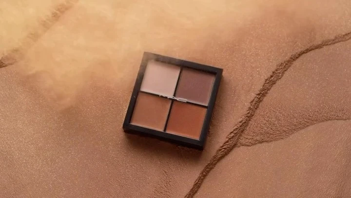 Close-up of a four-shade makeup palette, featuring neutral and earthy tones, displayed on a textured, shimmering bronze surface.