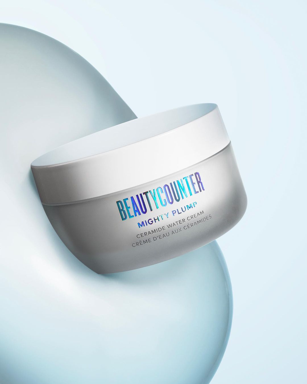 Beautycounter Mighty Plump Ceramide Water Cream jar against a light blue background with soft, rounded abstract forms.
