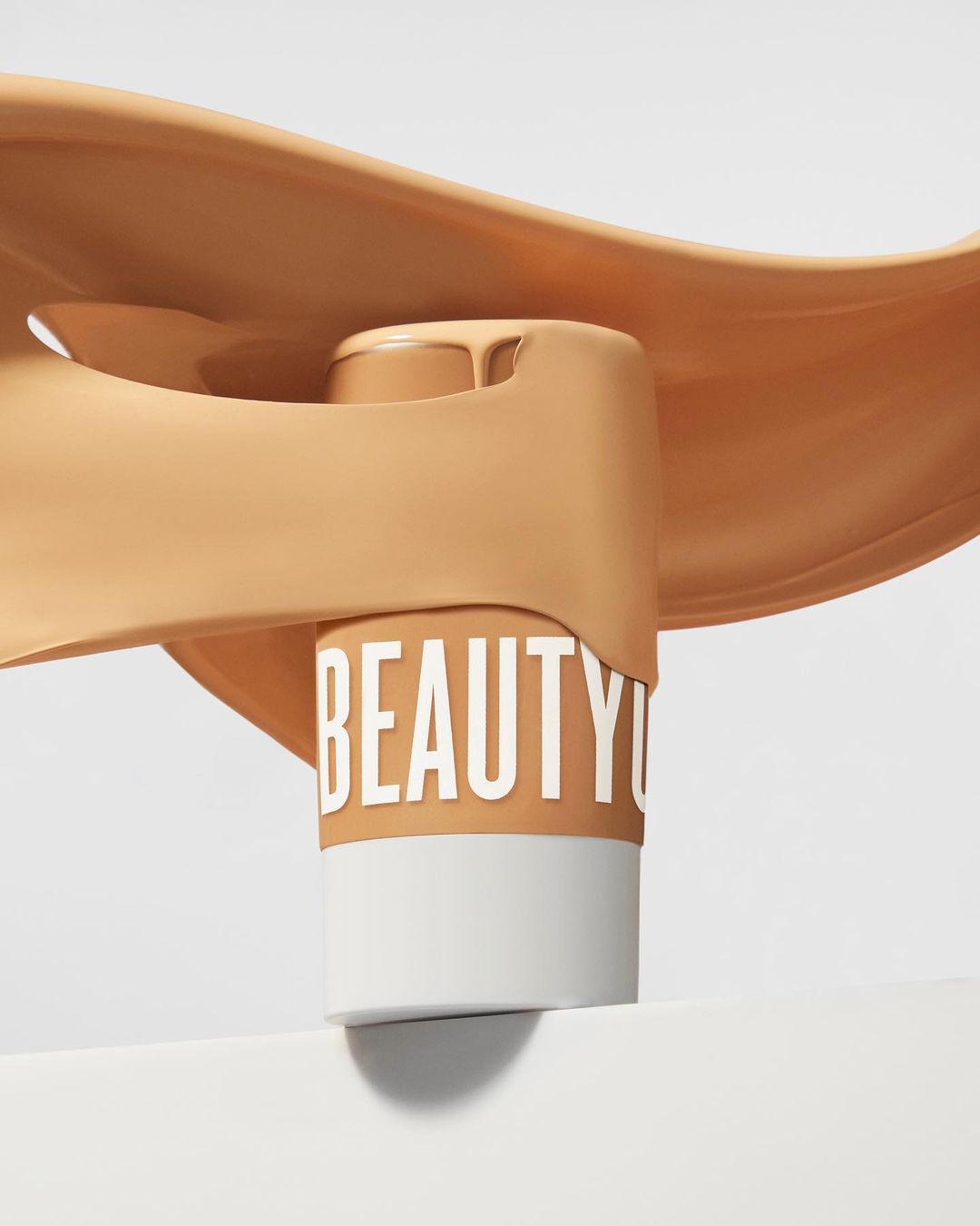 Tan foundation bottle with the word 'BEAUTY' around it, displayed with pouring liquid on white, for beauty and fashion promotion.